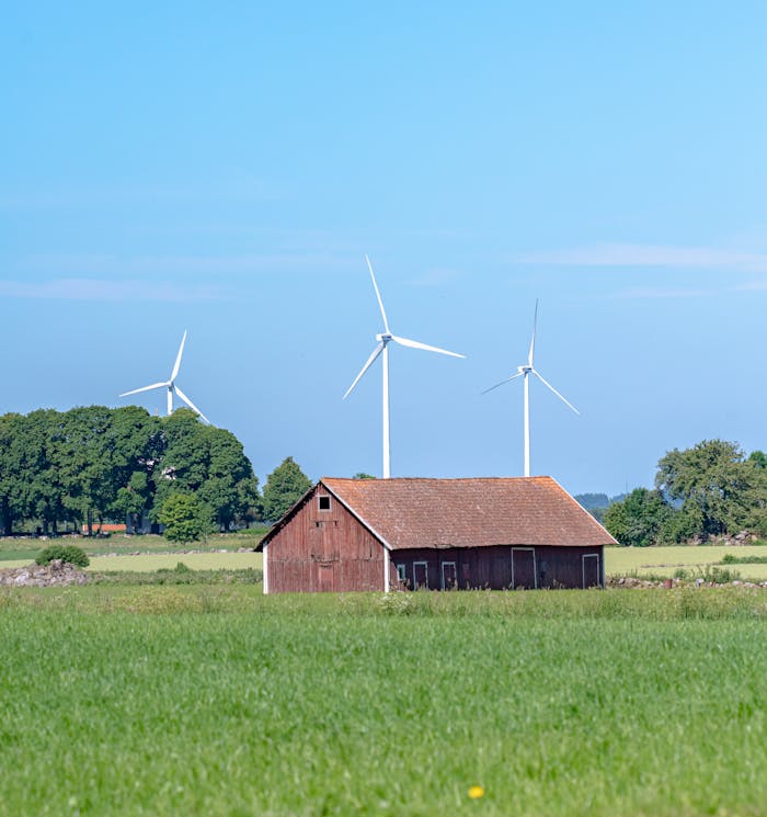 A red barn surrounded by green fields with wind turbines in the background, under a clear blue sky.